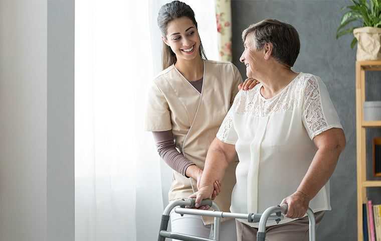 nurse in beige scrubs helping an elderly women using a walker in an assisted living facility